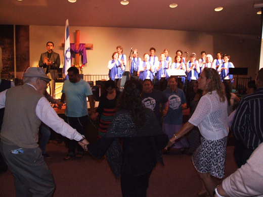 Jonathan Valverde at microphone besides Israeli flag and Beth Am Choir perform as a hora is danced by young and old, including 90-year-old Zimmerman